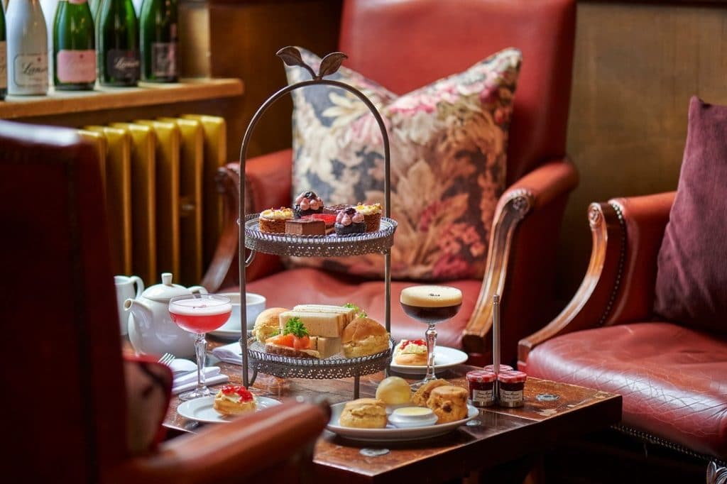 Assortment of afternoon tea items including cakes, sandwiches, scones and two cocktails on a table next to an armchair at the Hotel Du Vin