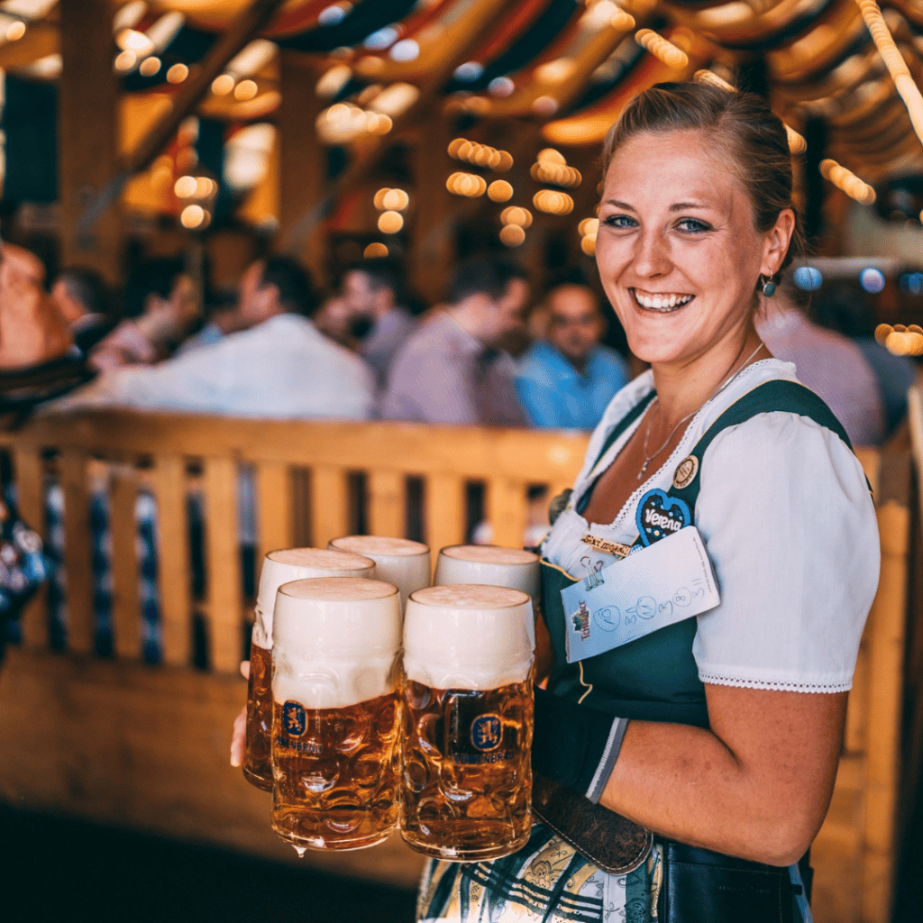 A woman holding five steins of beer and dressed in lederhosen