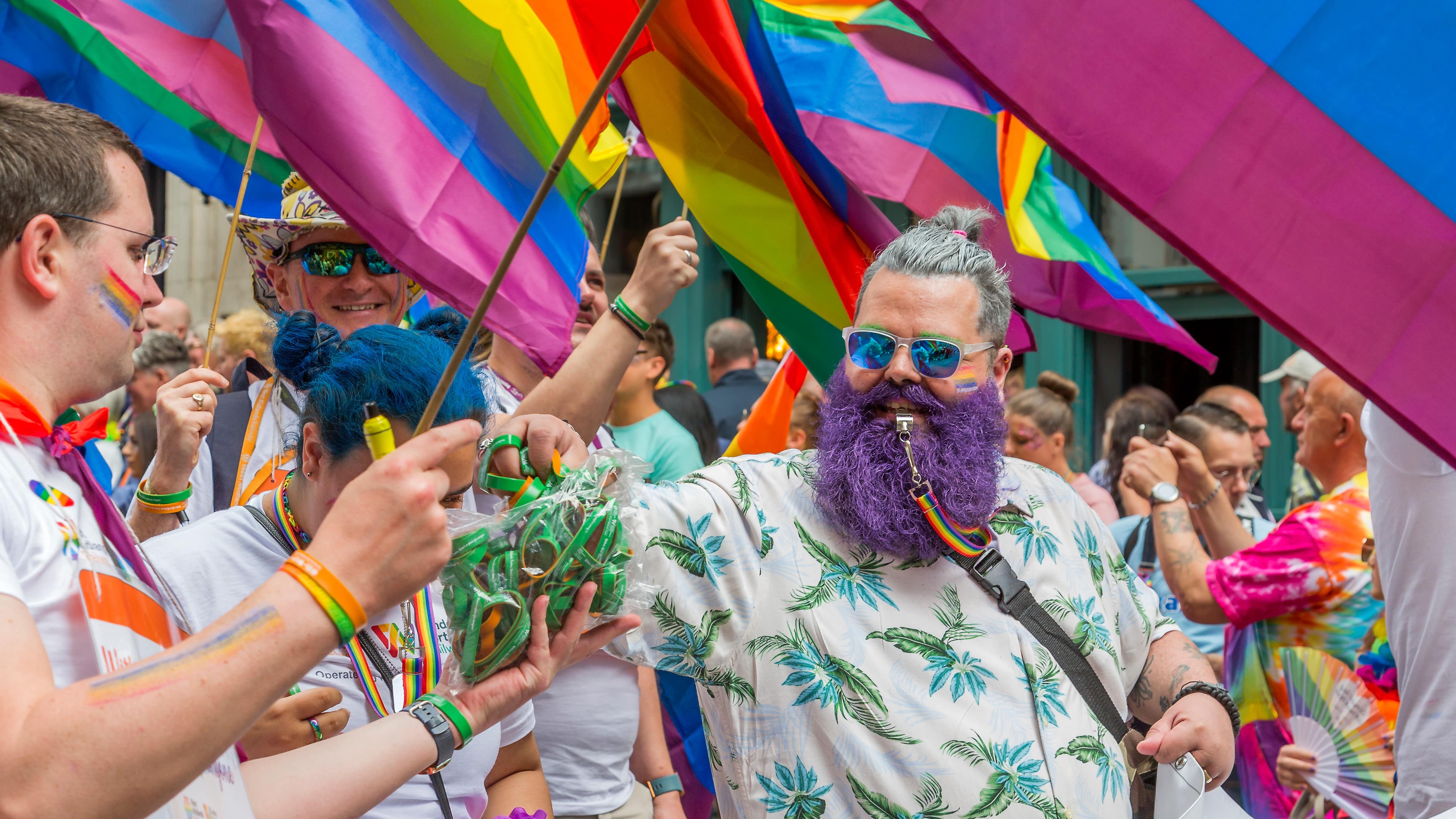 A reveller surrounded by flags and wearing a brightly coloured purple beard takes a handful of wristbands to hand out to the crowds along the Birmingham Pride 2023 parade route.