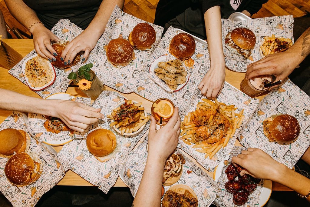 A table of fried chicken items from Yardbrds, with hand reaching for various items, such as burgers, fries and wings