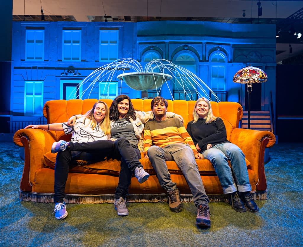 four people sit on the iconic orange Friends sofa in front of the fountain from the credits