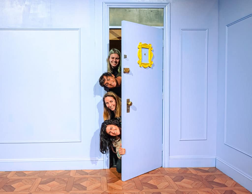 four people peek their heads around the purple door like the famous scene from Friends